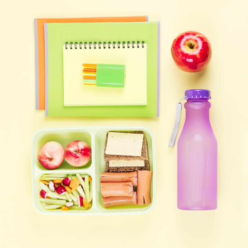 Imagen de una comida de picnic escolar con una fiambrera y una botella de agua reutilizable. Hay una libreta y lápices, una manzana y en la fiambrera, un sandwitch, fruta y ensalada. Sigue leyendo para conocer las mejores botellas de agua para niños.